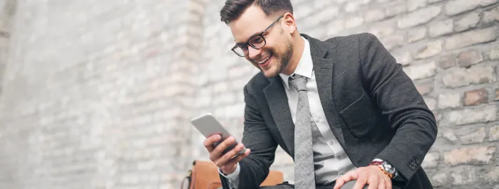 Uśmiechnięty mężczyzna w garniturze przegląda telefon, siedząc przy ceglanej ścianie